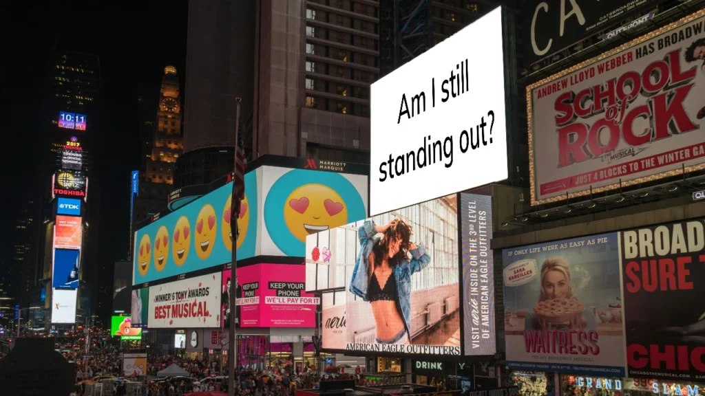 View of busy inner-city intersection with lots of billboards. One billboard reads "Am I still standing out?"